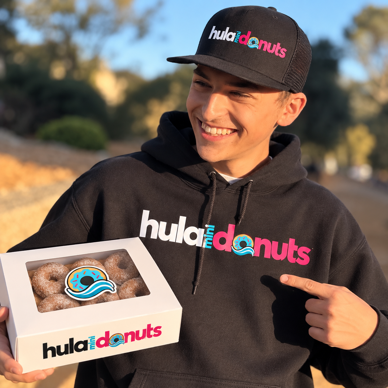 Smiling team member in a black Hula Mini Donuts hoodie and cap holding a box of cinnamon sugar mini donuts