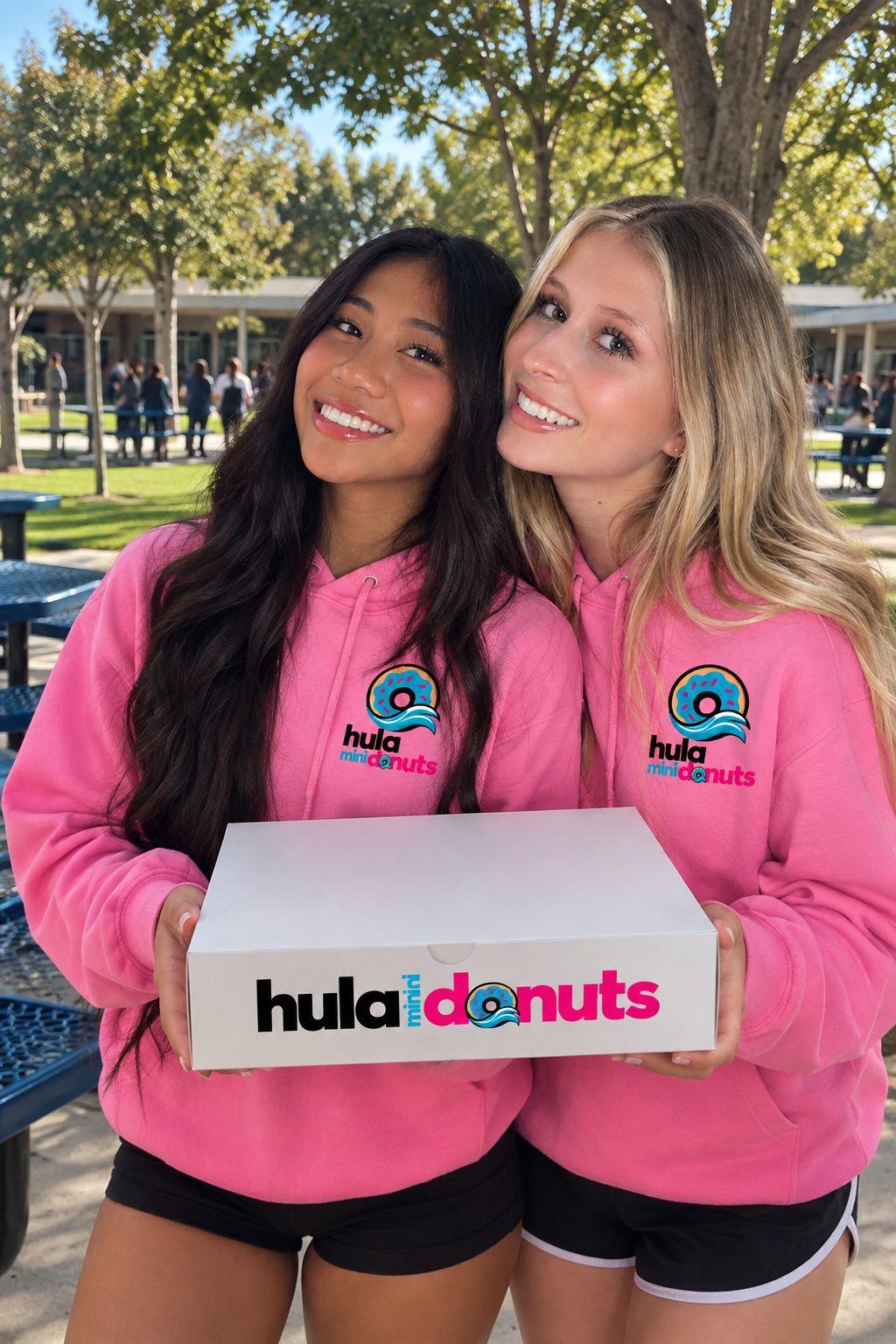 Two smiling team members in pink Hula Mini Donuts hoodies holding a big white donut box